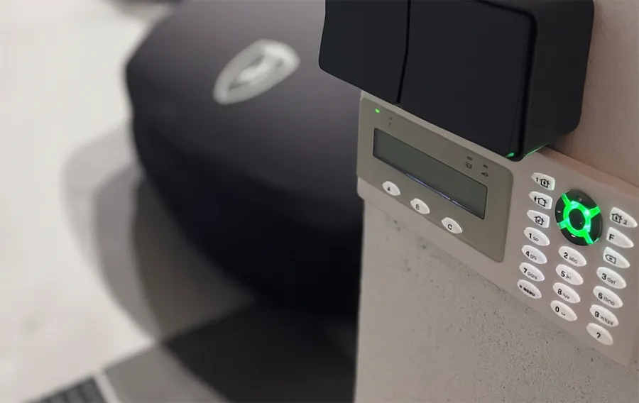 Clavier d'alarme de sécurité fixé au mur, doté de touches lumineuses et d'un petit écran, à côté d'un appareil noir.