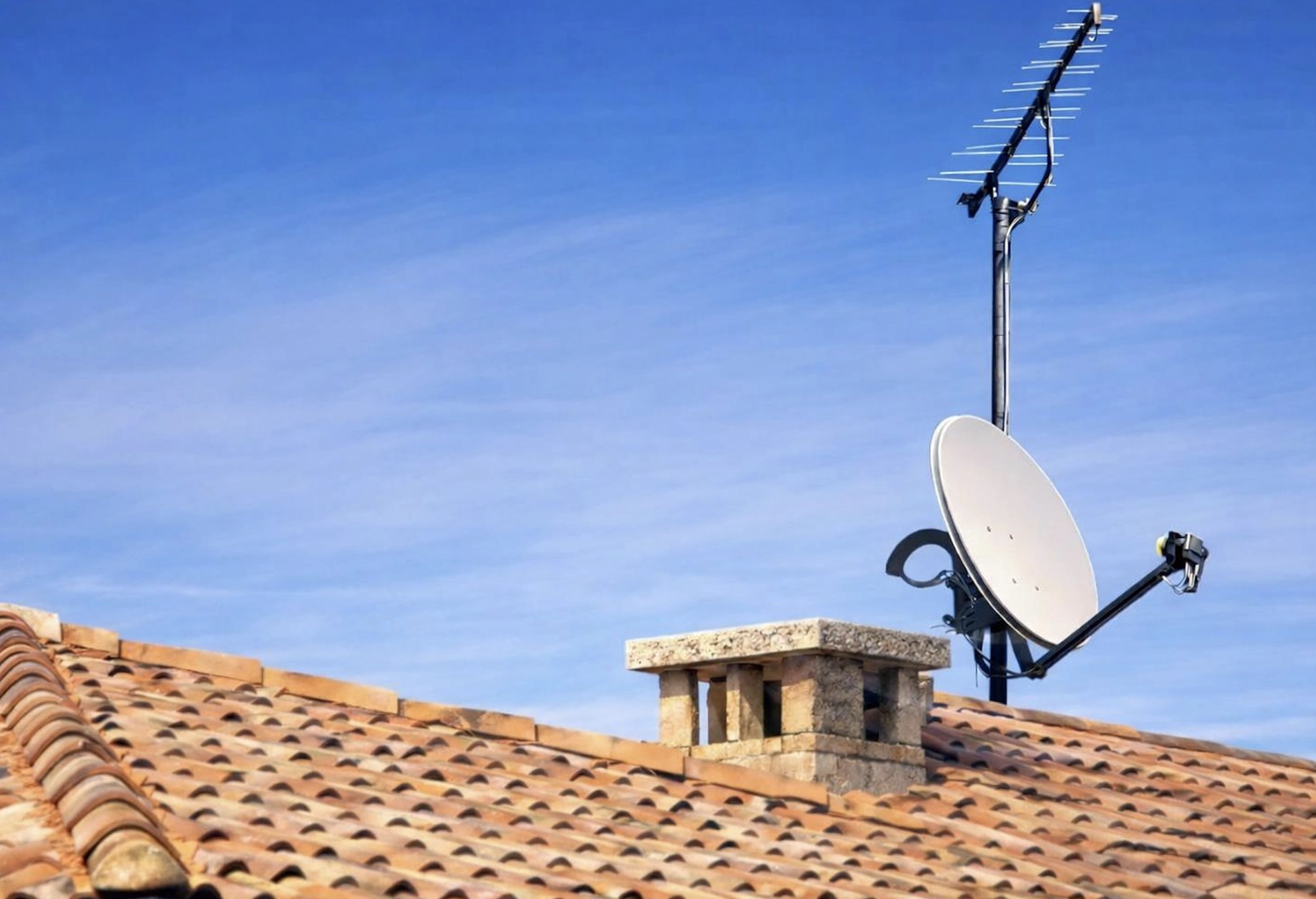 Toit en tuiles de terre cuite avec une cheminée en pierre, une antenne de télévision et une parabole, sur fond de ciel bleu clair.
