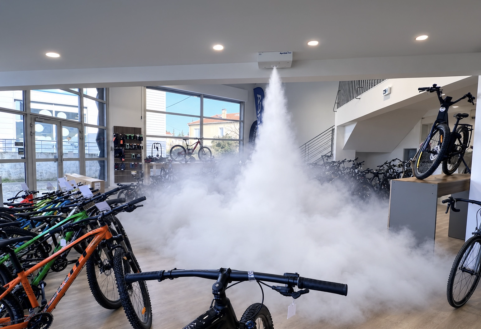 Intérieur d'un magasin de vélos où sont exposés plusieurs vélos, avec du brouillard ou de la brume s'échappant d'un dispositif fixé au plafond, au centre de la pièce.