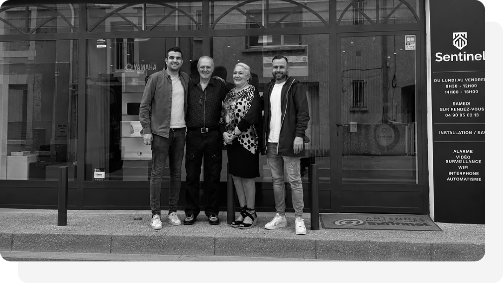 Pierre et Guillaume Sbaiz avec leur parent debout, bras dessus bras dessous, devant un bâtiment doté de portes vitrées et d'une enseigne « Sentinel ».
