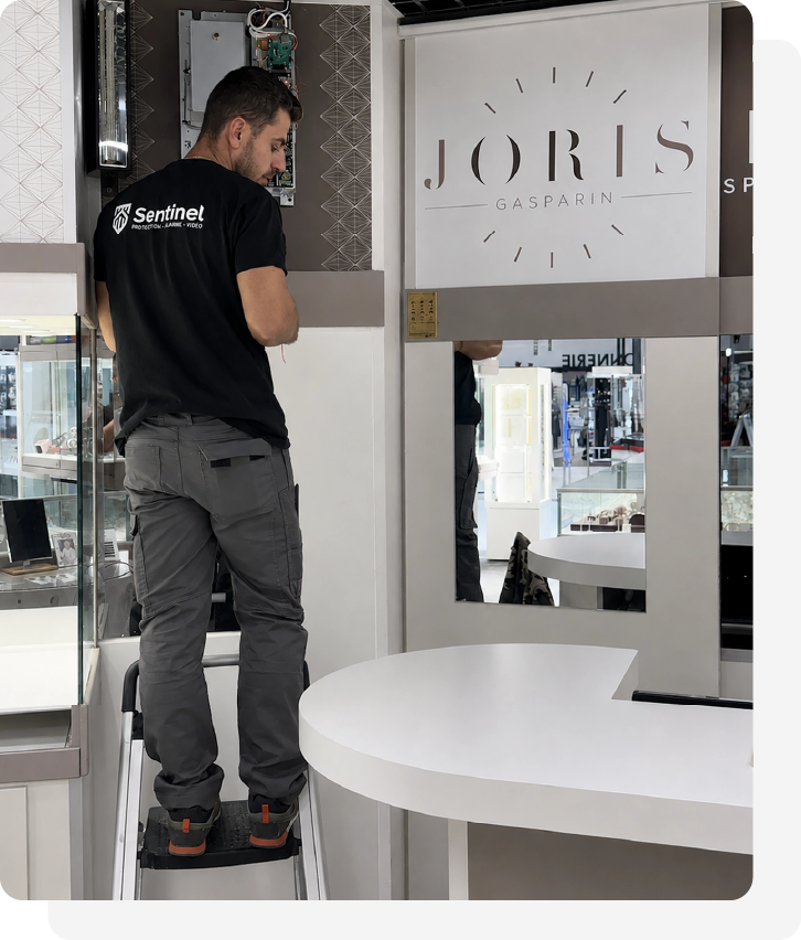 Un homme vêtu d'un t-shirt noir « Sentinel » se tient sur un escabeau et travaille sur un tableau électrique à l'intérieur d'un magasin moderne doté de vitrines en verre et d'un comptoir blanc incurvé.