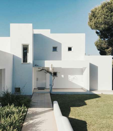 Maison blanche moderne à l'architecture cubique minimaliste, dotée d'un porche, de petites fenêtres et d'une allée dallée menant à l'entrée, le tout entouré de pelouse et d'arbres.