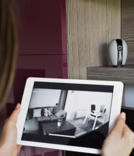 Gros plan sur une personne tenant une tablette qui affiche un flux vidéo en direct, en noir et blanc, d'un salon comprenant un canapé, des coussins, une table basse et une petite chaise sur laquelle est posé un objet ; l'image provient probablement d'une caméra de surveillance domestique.