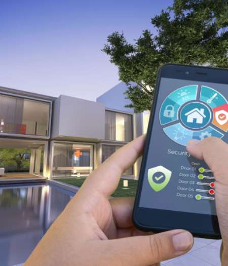 Une personne tient un smartphone affichant l'interface d'une application de sécurité pour maison connectée, devant une maison moderne dotée de grandes baies vitrées et d'une piscine, au crépuscule.