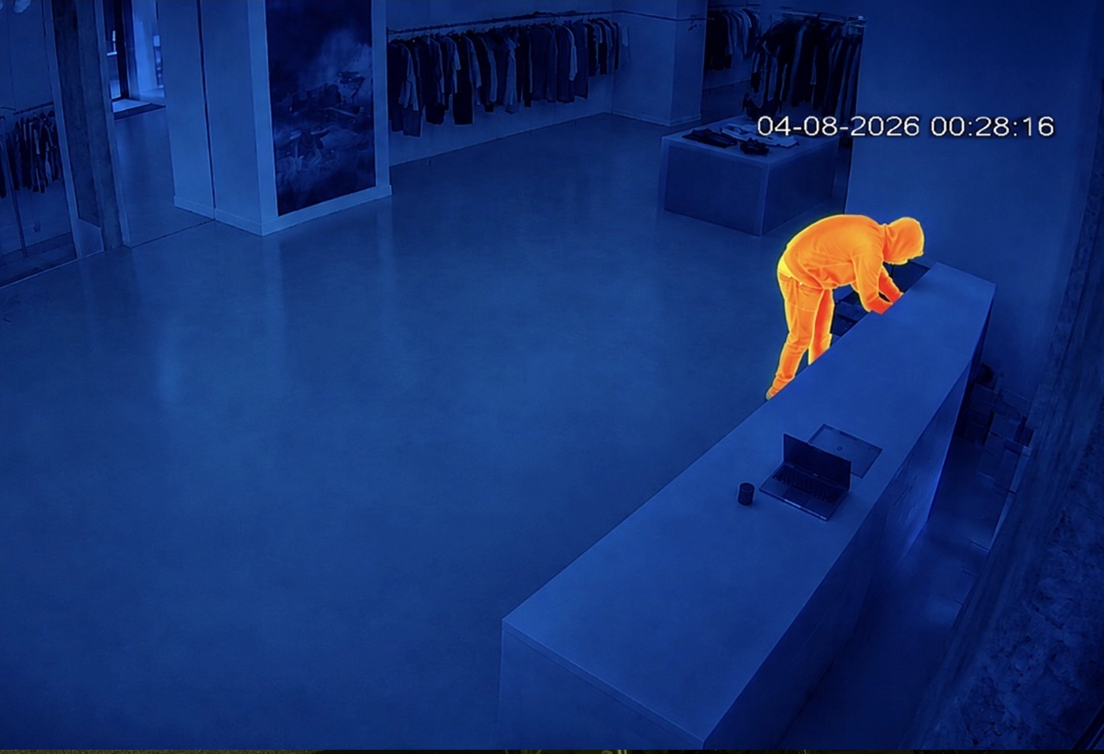 Image de caméra thermique montrant une personne portant une veste à capuche à l'intérieur d'un magasin de vêtements, penchée sur un comptoir où se trouvent un ordinateur portable et d'autres objets, avec l'horodatage 04-08-2026 00:28:16.