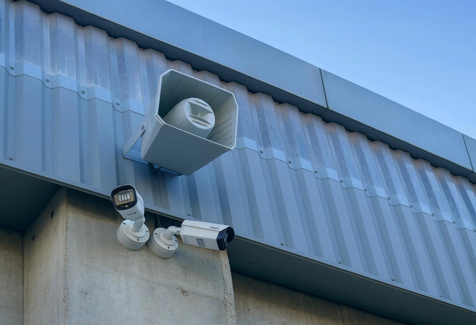 Deux caméras de surveillance blanches et un grand haut-parleur d'extérieur blanc fixés à l'angle d'un bâtiment en béton au bardage métallique ondulé, sous un ciel bleu dégagé.