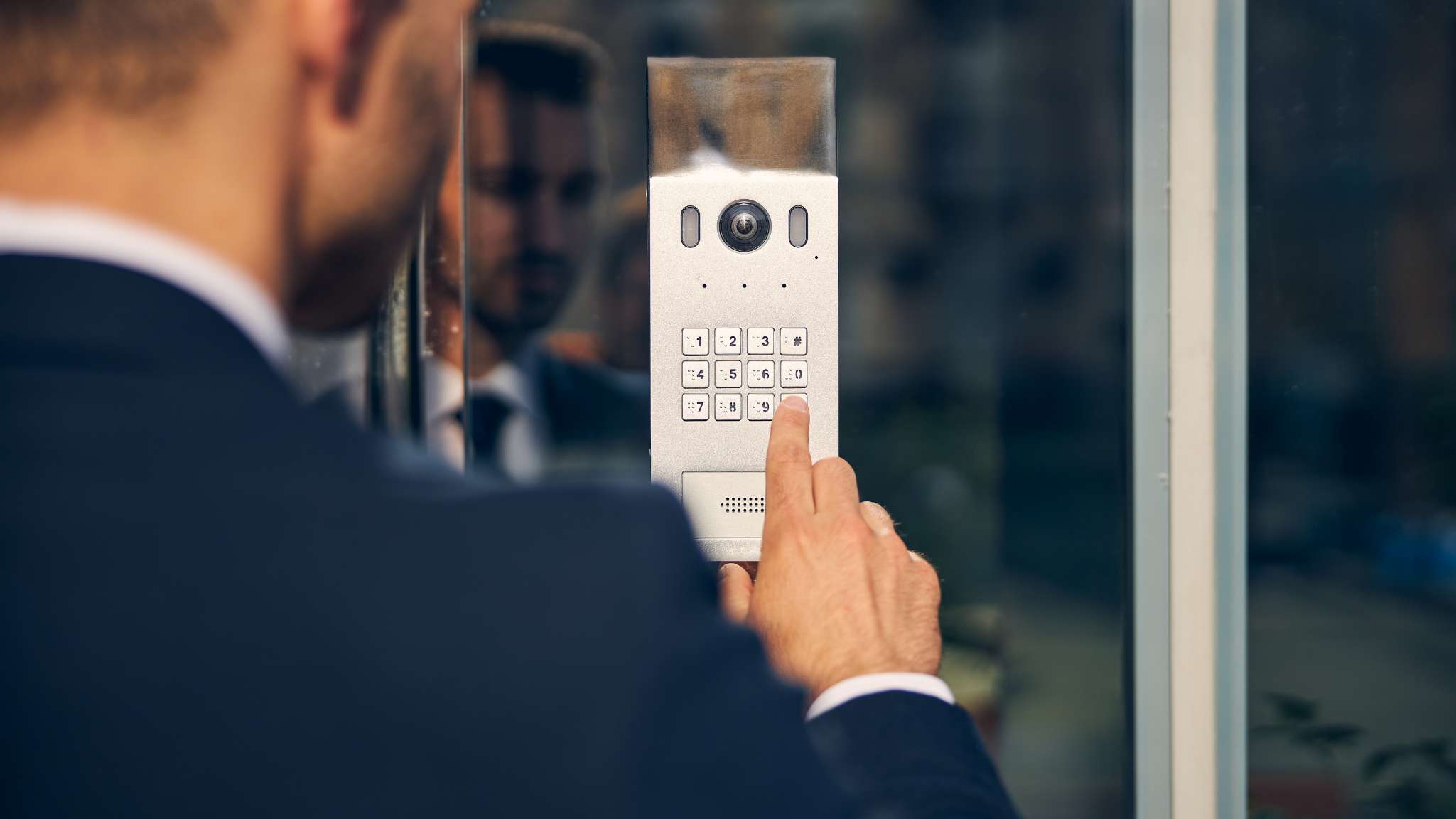 Un homme en costume appuie sur une touche du clavier d'un interphone argenté fixé sur une porte vitrée, son reflet étant visible dans la vitre.