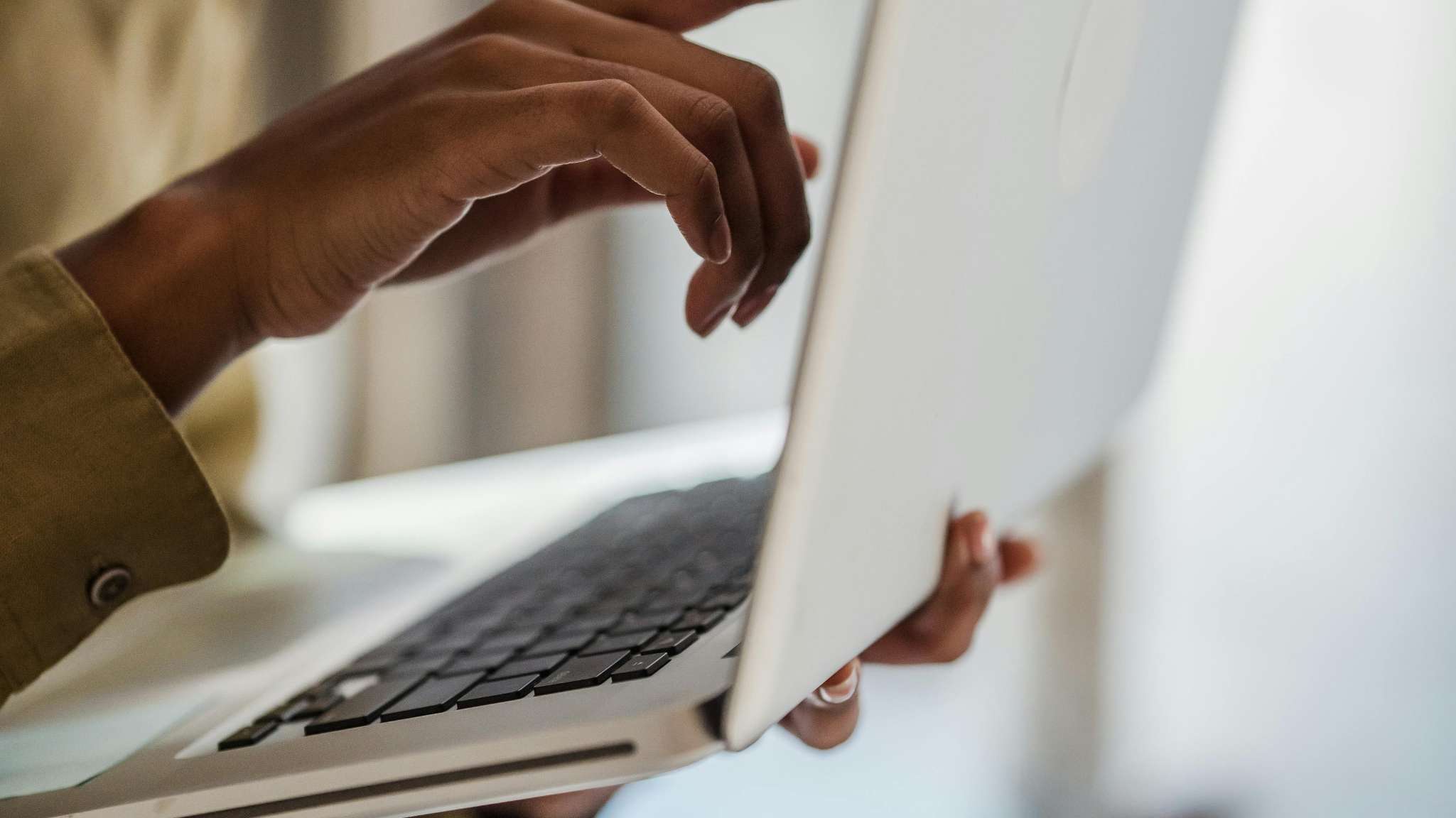 Gros plan sur les mains à la peau foncée d'une personne tenant un ordinateur portable argenté au clavier noir ; une main soutient l'appareil par-dessous tandis que l'autre est positionnée au-dessus des touches.