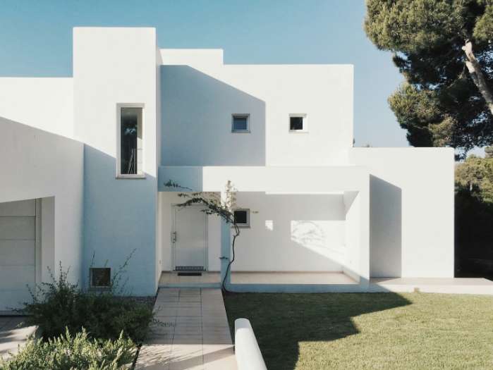 Maison blanche moderne à l'architecture cubique minimaliste, dotée d'un porche, de petites fenêtres et d'une allée dallée menant à l'entrée, le tout entouré de pelouse et d'arbres.
