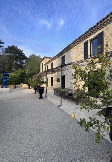 Bâtiment en pierre avec toit en tuiles et volets noirs, bordant une cour gravillonnée agrémentée de tables d'extérieur, de plantes en pot et d'arbres, sous un ciel bleu partiellement nuageux.
